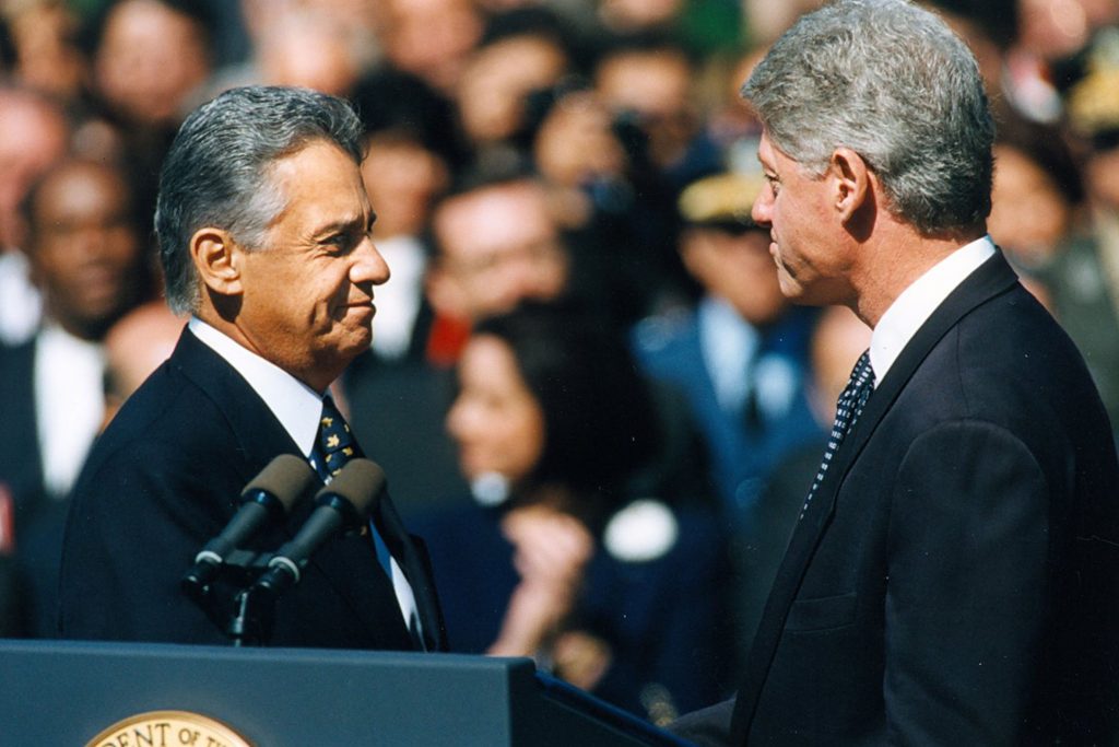 Fotografia colorida de um encontro entre Fernando Henrique Cardoso, à esquerda, e Bill Clinton, à direita, nos Estados Unidos. À frente deles, um púlpito com dois microfones.