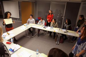 Fotografia colorida de um evento do Acervo em uma sala da Fundação FHC. Em volta de uma mesa, sentadas e em pé, pessoas assistem à palestra de uma mulher. Ela está em pé e segura, com as duas mãos, um papel.