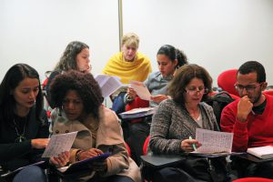 Fotografia colorida de pessoas no auditório da Fundação FHC. Elas estão sentadas. São participantes da oficina do Acervo. Em pequenos grupos, estão analisando alguns documentos.