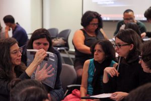 Fotografia colorida de uma oficina do Acervo no auditório da Fundação FHC. À esquerda, a palestrante. Ela segura um espécie de saquinho transparente com as mãos; está falando. À direita, sentadas, pessoas que estão participando do evento. Elas estão olhando para a explicação da palestrante.