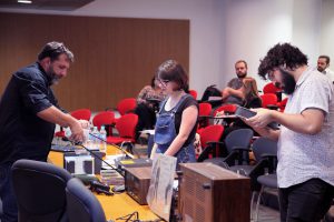 Fotografia colorida de uma oficina do Acervo no auditório da Fundação FHC. À esquerda, o palestrante. Ele está em pé, veste uma camisa e está atrás de uma mesa de madeira. Segura, com as mãos, objetos do Acervo. À frente delem duas pessoas que estão participando do evento.