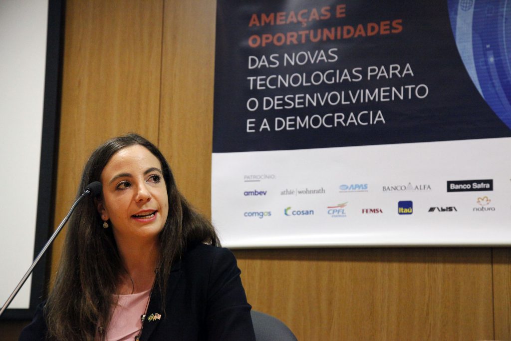 Fotografia colorida de um evento na Fundação FHC. À esquerda, sentada, a palestrante Lindsay. Ele veste terno preto e blusa rosa. Ela está falando em um microfone que está sobre a mesa à frente. Atrás dela, um banner do evento em um painel de madeira.