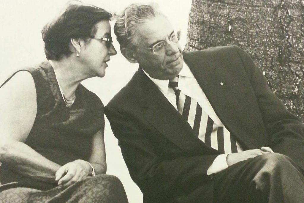 Fotografia em preto e branco de Ruth Cardoso e Fernando Henrique Cardoso. Eles estão sentados. Ruth está falando ao pé do ouvido de FHC.