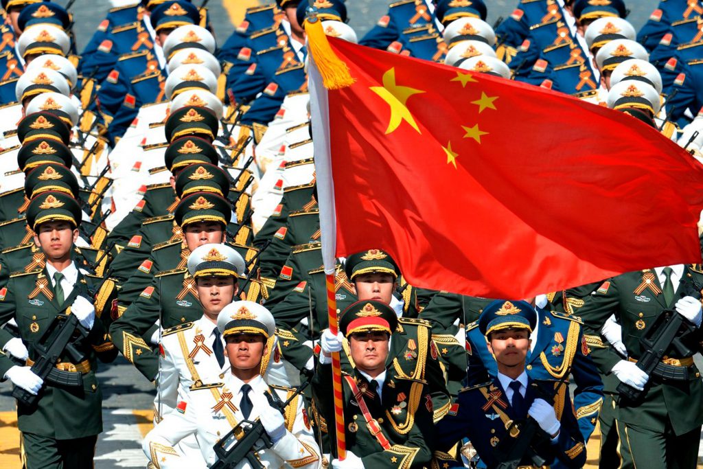 Fotografia colorida de militares chineses em um desfile. Um dos militares segura, com as duas mãos, a bandeira da China em uma haste.