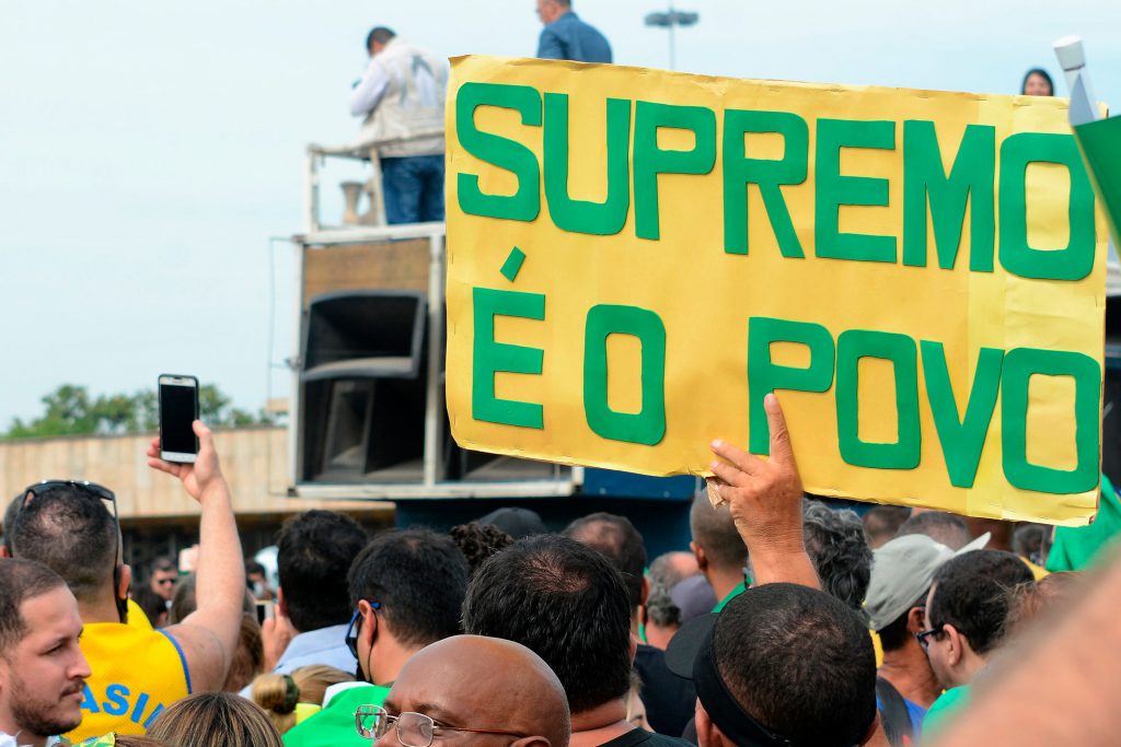 Fotografia colorida que mostra pessoas em uma manifestação. Em destaque na foto, uma pessoa segura um cartaz amarelo com a seguinte frase, escrita em verde: Supremo é o Povo.