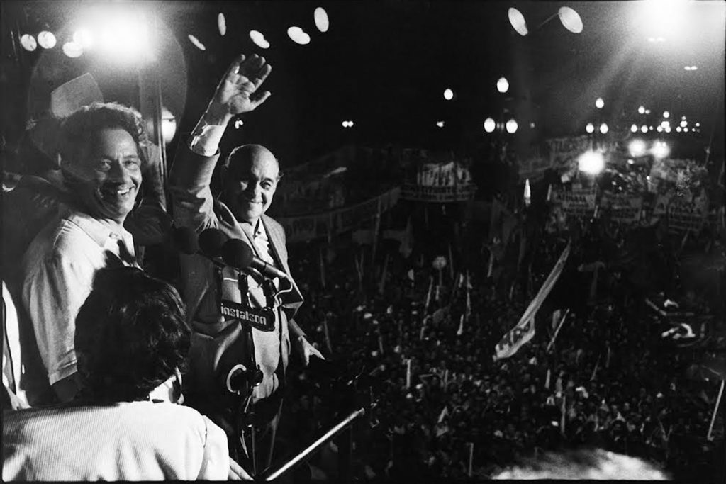 Fotografia em preto e branco de um comício nos anos 1980. À esquerda estão Fernando Henrique Cardoso e Tancredo Neves. Ambos estão olhando para a câmera e sorrindo. Eles estão em uma espécie de palanque. Ao fundo, uma multidão de pessoas.