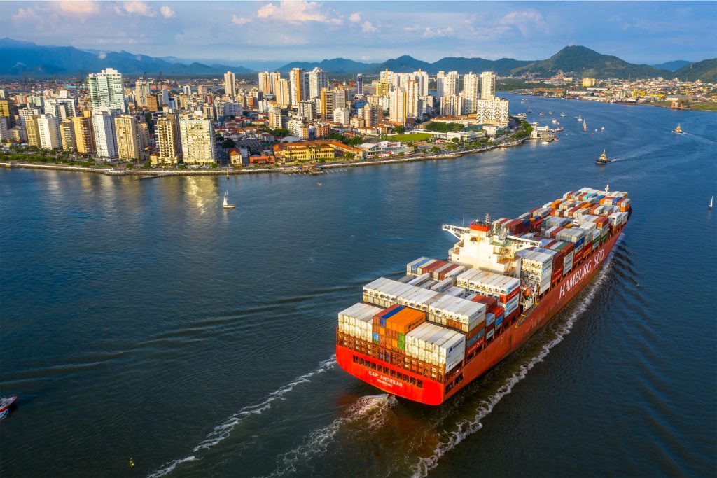 Fotografia colorida e aérea que mostra, à direita, um navio com diversos contêineres navegando por um rio. À esquerda, mais ao fundo, uma cidade.