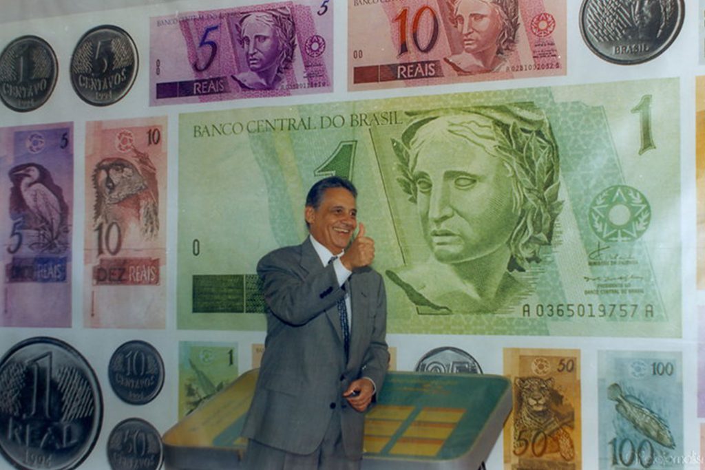 Fotografia colorida de Fernando Henrique Cardoso em frente a um painel com cédulas de real. Em destaque, a nota de um real. FHC veste terno, camisa social e gravata. Com a mão direita, ele faz um gesto de positivo.