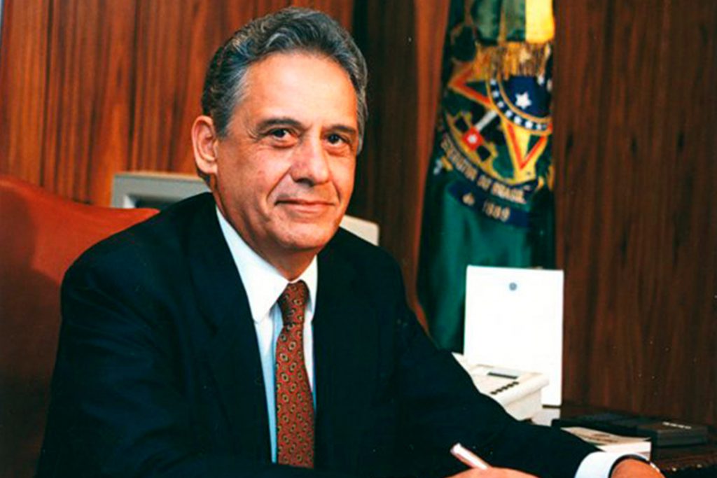 Fotografia colorida de Fernando Henrique Cardoso no primeiro dia como presidente do Brasil. Ele está sentado. Veste terno, camisa social e gravata. Sorri levemente. Ao fundo, em um mastro, a bandeira da República.