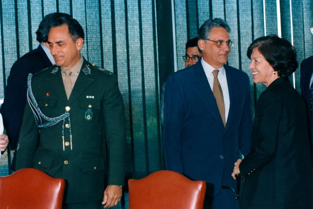 Fotografia colorida de um evento no Palácio do Planalto, em Brasília. Uma mulher e dois homens aparecem na foto. Todos estão em pé. Um dos homens é Fernando Henrique Cardoso, que veste terno, camisa social, calça e gravata. Usa óculos. O outro homem é um militar do Exército. A mulher é Eunice Paiva.