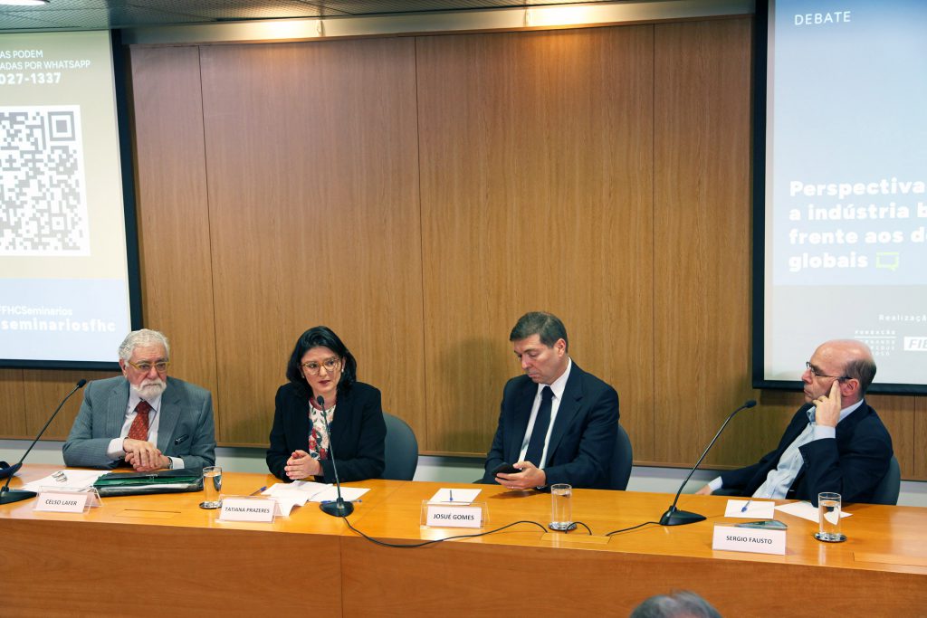 Fotografia colorida que mostra os quatro participantes do evento sentados atrás de uma mesa de madeira. Da esquerda para a direita, são eles: Celso Lafer, Tatiana Prazeres, que está falando, Josué Gomes e Sergio Fausto. Os três homens vestem terno. Tatiana veste uma blusa branca e um terninho preto por cima. Ao fundo, painel de madeira.