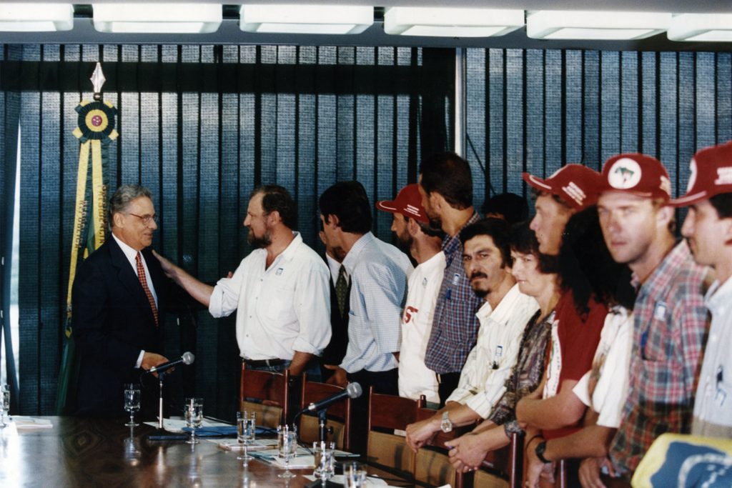 Fotografia colorida de um encontro de Fernando Henrique Cardoso, à esquerda, com membros do MST, à direita. A maioria deles usa um boné vermelho na cabeça. FHC veste terno, camisa social, calça e gravata. Ele está cumprimentando as pessoas.