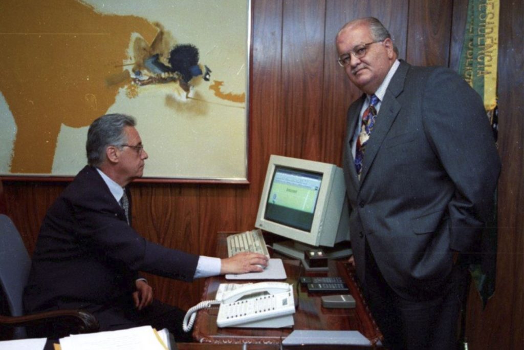 Fotografia colorida de Fernando Henrique Cardoso com Sergio Motta, ministro das Comunicações. FHC está sentado à frente de um computador. Ele está olhando para a tela. Sergio Motta está olhando para a câmera. Ambos vestem terno, camisa social e gravata.