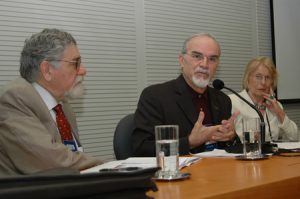 Fotografia colorida de um evento do Acervo no auditório da Fundação FHC. Sentados atrás de uma mesa de madeira, três palestrantes, dois homens e uma mulher. O homem que está ao centro está falando.