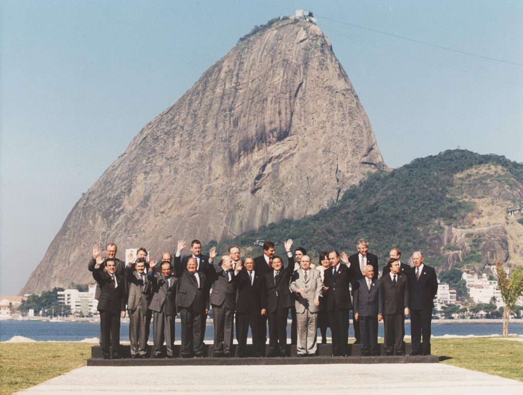 Fotografia colorida de uma reunião com presidentes da América Latina no Rio de Janeiro. Em primeiro plano, os chefes de Estado. Eles estão em pé e acenam com as mãos. Ao fundo, o Pão de Açúcar.