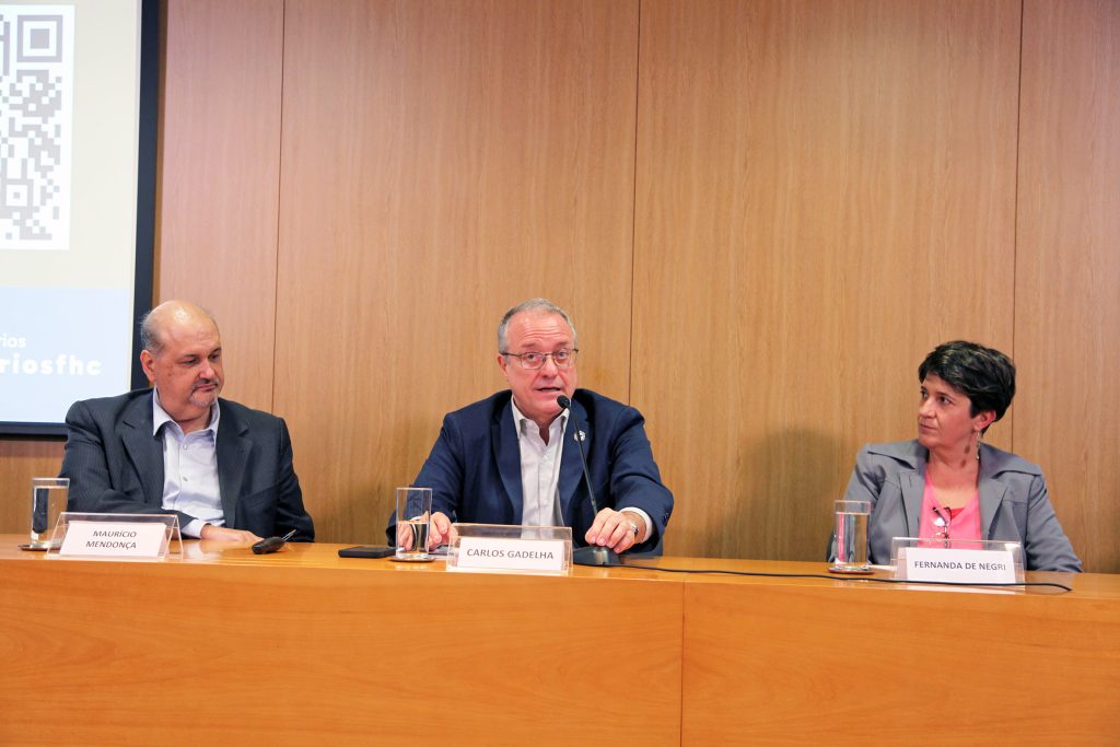 Fotografia colorida de um evento no auditório da Fundação FHC. Dois homens e uma mulher estão sentados atrás de uma mesa de madeira. O homem que está ao centro está falando; há um microfone sobre a mesa.