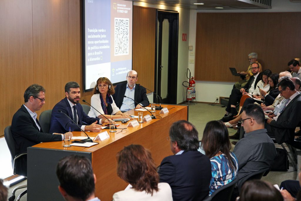 Fotografia colorida de um evento no auditório da Fundação FHC. À direita, a plateia. À esquerda, os palestrantes. Todos estão sentados atrás de uma mesa de madeira.