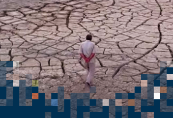 Fotografia colorida que mostra um homem andando em um terreno extremamente seco, com diversas ranhuras no solo, sinal de que não há chuva. O homem veste uma roupa branca e está com as mãos para trás.