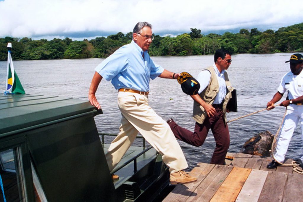 Fotografia colorida que mostra Fernando Henrique Cardoso saindo de uma embarcação que está na beira de um rio. FHC veste camisa azul, calça bege e sapato. Ele segura, com a mão esquerda, um boné. Ele está saltando para um píer que está à frente.