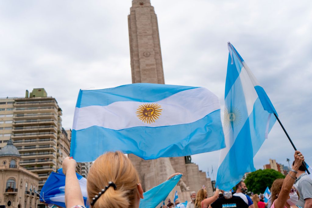 Fotografia colorida de uma manifestação na Argentina. Em primeiro plano, ao centro, uma bandeira do país vizinho, tremulando em uma haste. Ao fundo, um monumento histórico.