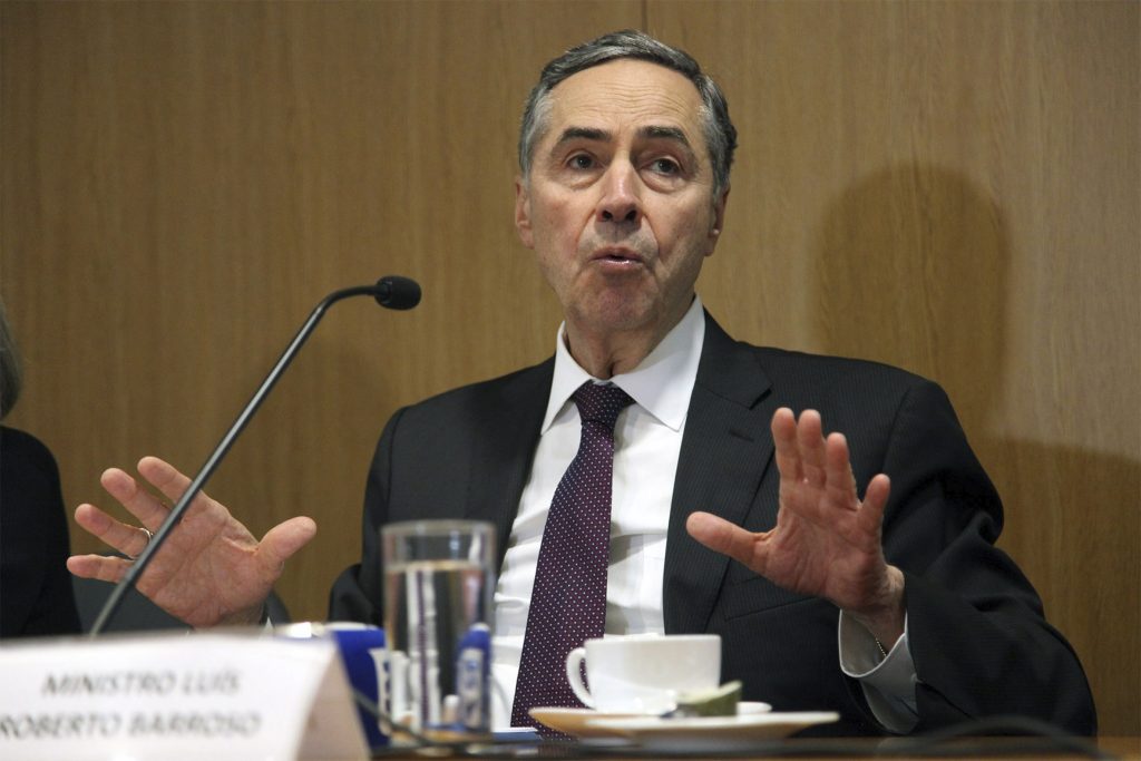 Fotografia colorida do ministro Luís Roberto Barroso em um debate no auditório da Fundação FHC. Ele está sentado, veste terno preto, camisa social branca e gravata. Está falando e gesticulando com as duas mãos. À frente dele, sobre a mesa, um microfone. Ao fundo, painel de madeira.