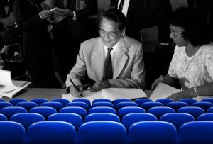 Fotografia em preto e branco de Fernando Henrique Cardoso assinando um documento que está sobre uma mesa, à frente dele. Ao lado de FHC, uma mulher observa a cena. Em primeiro plano, em azul, poltronas que lembram as de um cinema.