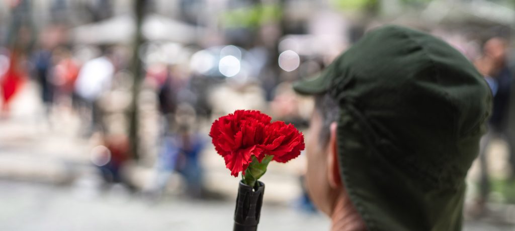 Fotografia colorida de um militar de costas. À frente dele, uma arma que tem, na ponta do cano, um cravo vermelho. Ao fundo, desfocadas, pessoas andando.