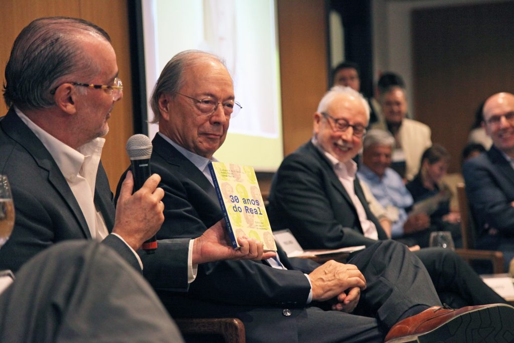 Fotografia colorida de um debate sobre o Plano Real na Fundação FHC. A foto mostra os palestrantes, todos sentados. Um deles está falando. Além do microfone, ele segura e mostra um livro sobre os 30 anos do Plano Real. Os outros palestrantes estão ao lado dele, também sentados. Eles estão sorrindo.