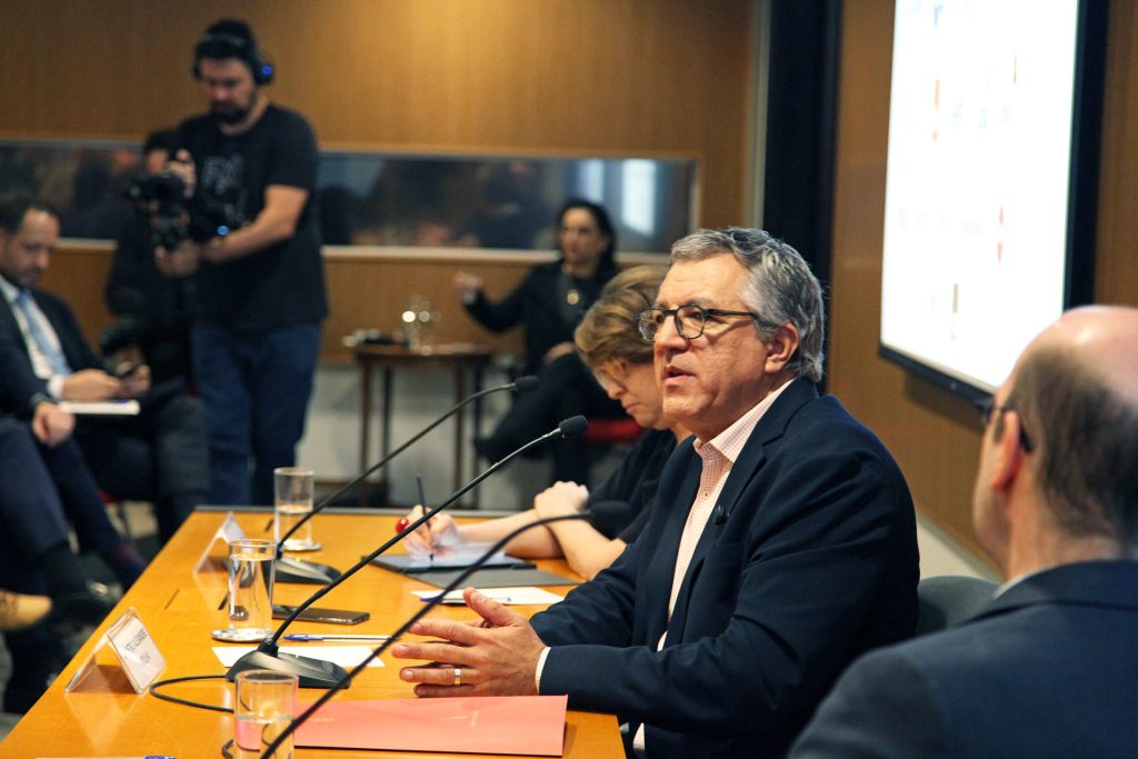 Fotografia colorida de um debate no auditório da Fundação FHC. À direita, atrás de uma mesa de madeira, os palestrantes. Todos estão sentados. Sobre a mesa, microfones. Um homem está falando. Ele olha para a plateia, que está à frente.