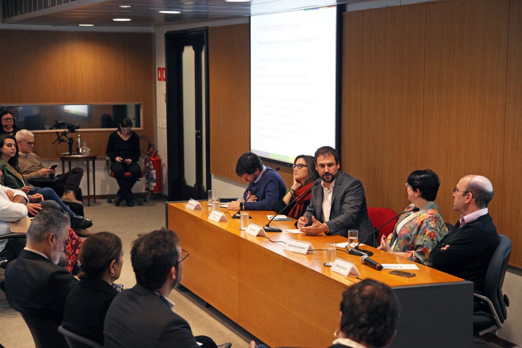 Fotografia colorida de um debate no auditório da Fundação FHC. À direita, sentados atrás de uma mesa de madeira, os palestrantes. Um deles está falando. À esquerda, pessoas na plateia.