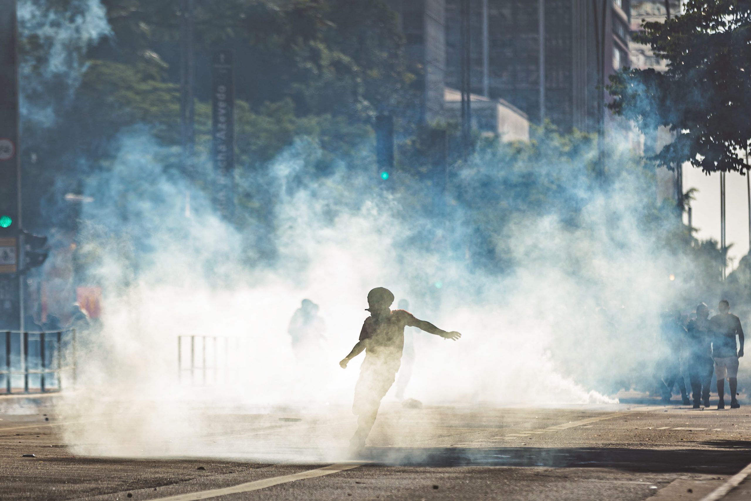 Fotografia colorida de pessoas em uma manifestação na Avenida Paulista, em São Paulo. Ao centro, uma pessoa está com o corpo inclinado, como se fosse chutar um objeto que está no chão. Em volta da pessoa, bastante fumaça.