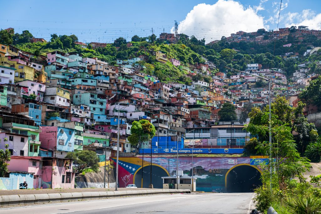 Fotografia colorida de uma cidade na Venezuela. Em primeiro plano, uma rua que leva até um túnel. Ao fundo e sobre o túnel, diversas casas coloridas em um morro.