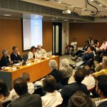 Fotografia colorida de um debate na Fundação FHC. À direita, a plateia. Todas as pessoas estão sentadas. À esquerda, sentados atrás de uma mesa de madeira, os palestrantes. Uma mulher está falando. Sobre a mesa, um microfone.