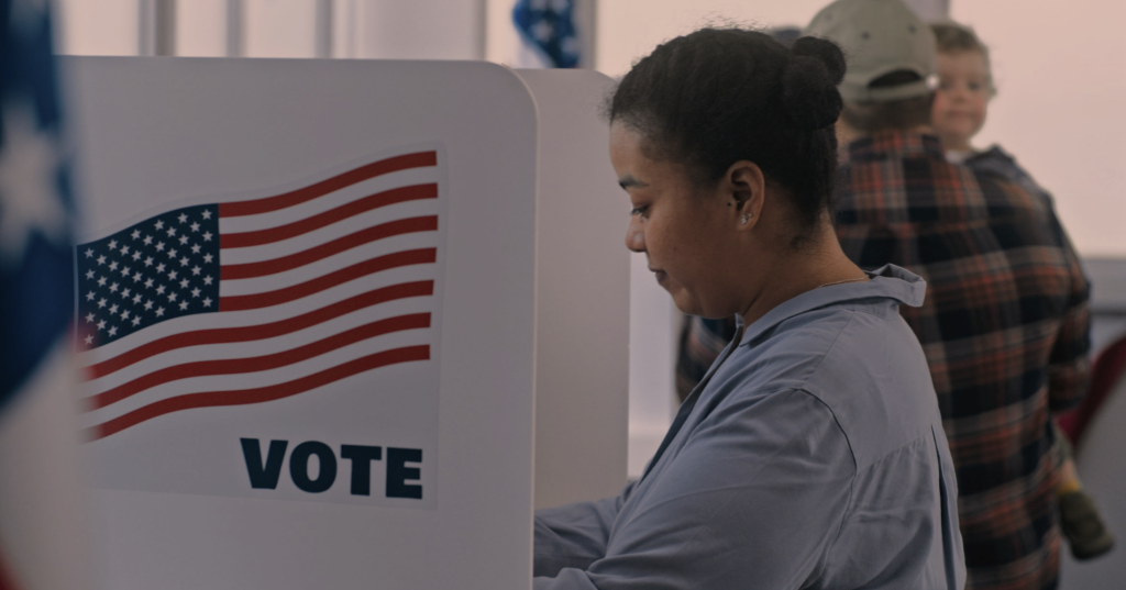 Fotografia colorida que mostra uma mulher em um ambiente de votação nos Estados Unidos. À esquerda, a cabine de votação, com a bandeira dos Estados Unidos e a palavra 