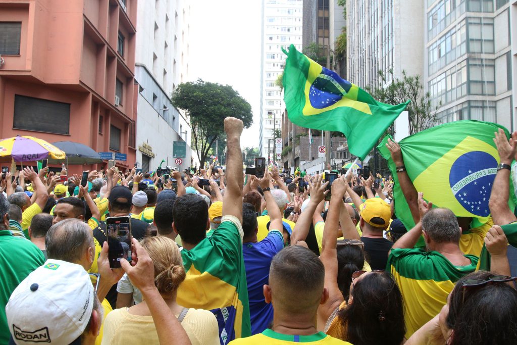 Fotografia colorida de pessoas em uma manifestação na cidade de São Paulo. Muitas pessoas vestem a camisa verde e amarela da Seleção Brasileira de Futebol. Há também algumas bandeiras do Brasil tremulando.