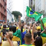 Fotografia colorida de pessoas em uma manifestação na cidade de São Paulo. Muitas pessoas vestem a camisa verde e amarela da Seleção Brasileira de Futebol. Há também algumas bandeiras do Brasil tremulando.