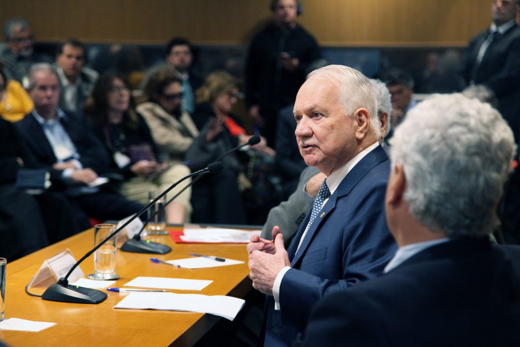 Fotografia colorida que mostra, ao centro, em destaque, o ministro Edson Fachin. Ele está sentado atrás de uma mesa de madeira. À frente dele, sobre a mesa, microfones, papéis e copos de água. Ele está falando e gesticulando com as mãos. Ao fundo, desfocadas, pessoas na plateia.