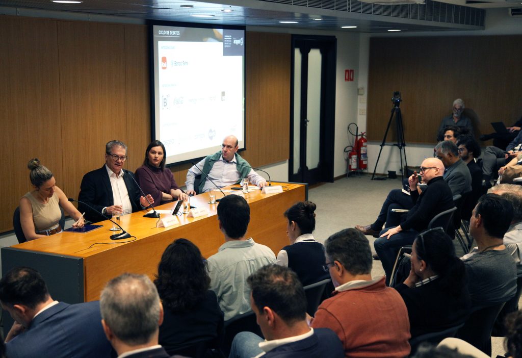 Fotografia colorida que mostra, à esquerda, sentados, atrás de uma mesa de madeira, os palestrantes em um debate na Fundação FHC. À direita, também sentadas, pessoas na plateia.