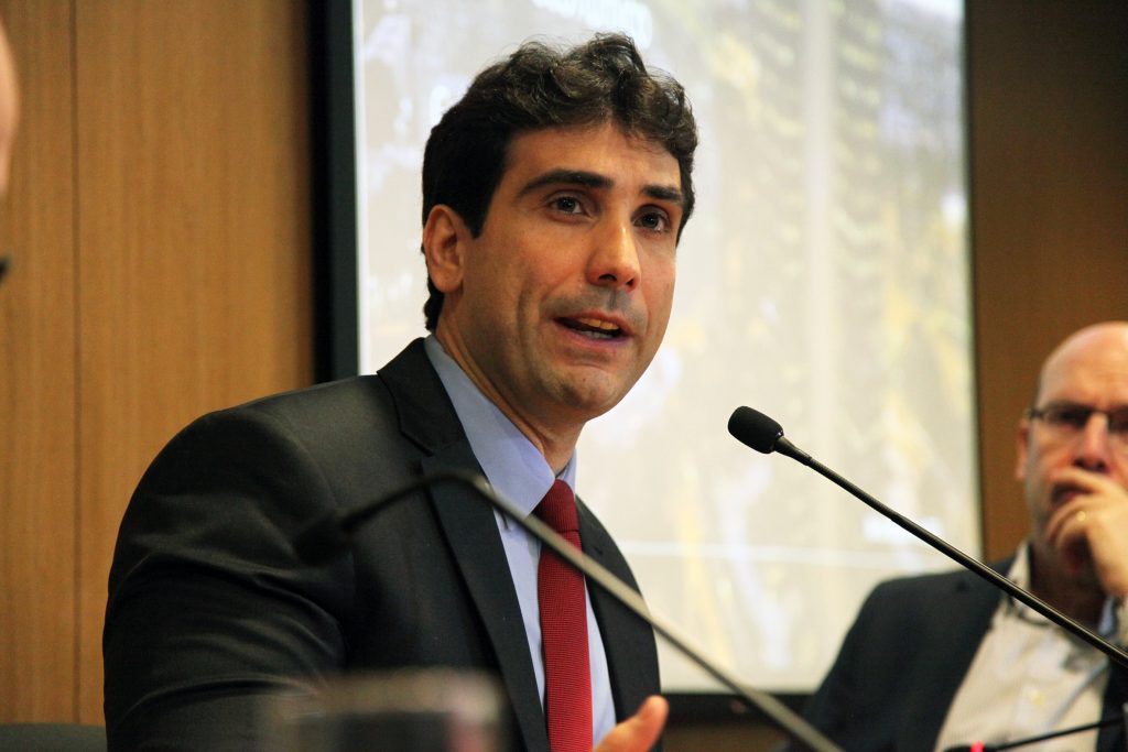 Fotografia colorida que mostra o economista e presidente do Banco Central Gabriel Galípolo em debate na Fundação FHC. Ele está sentado. Veste terno, camisa social azul e gravata vermelha. Galípolo está falando em um microfone que está à frente dele, sobre uma mesa de madeira.