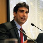 Fotografia colorida que mostra o economista e presidente do Banco Central Gabriel Galípolo em debate na Fundação FHC. Ele está sentado. Veste terno, camisa social azul e gravata vermelha. Galípolo está falando em um microfone que está à frente dele, sobre uma mesa de madeira.