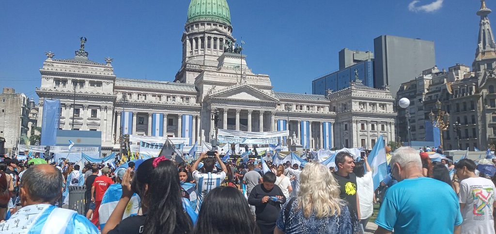 Fotografia colorida que mostra pessoas em uma manifestação em Buenos Aires, em frente ao Congresso. A maioria das pessoas usa camisa com cores azul e branco. Há também cartazes e bandeiras.