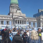 Fotografia colorida que mostra pessoas em uma manifestação em Buenos Aires, em frente ao Congresso. A maioria das pessoas usa camisa com cores azul e branco. Há também cartazes e bandeiras.