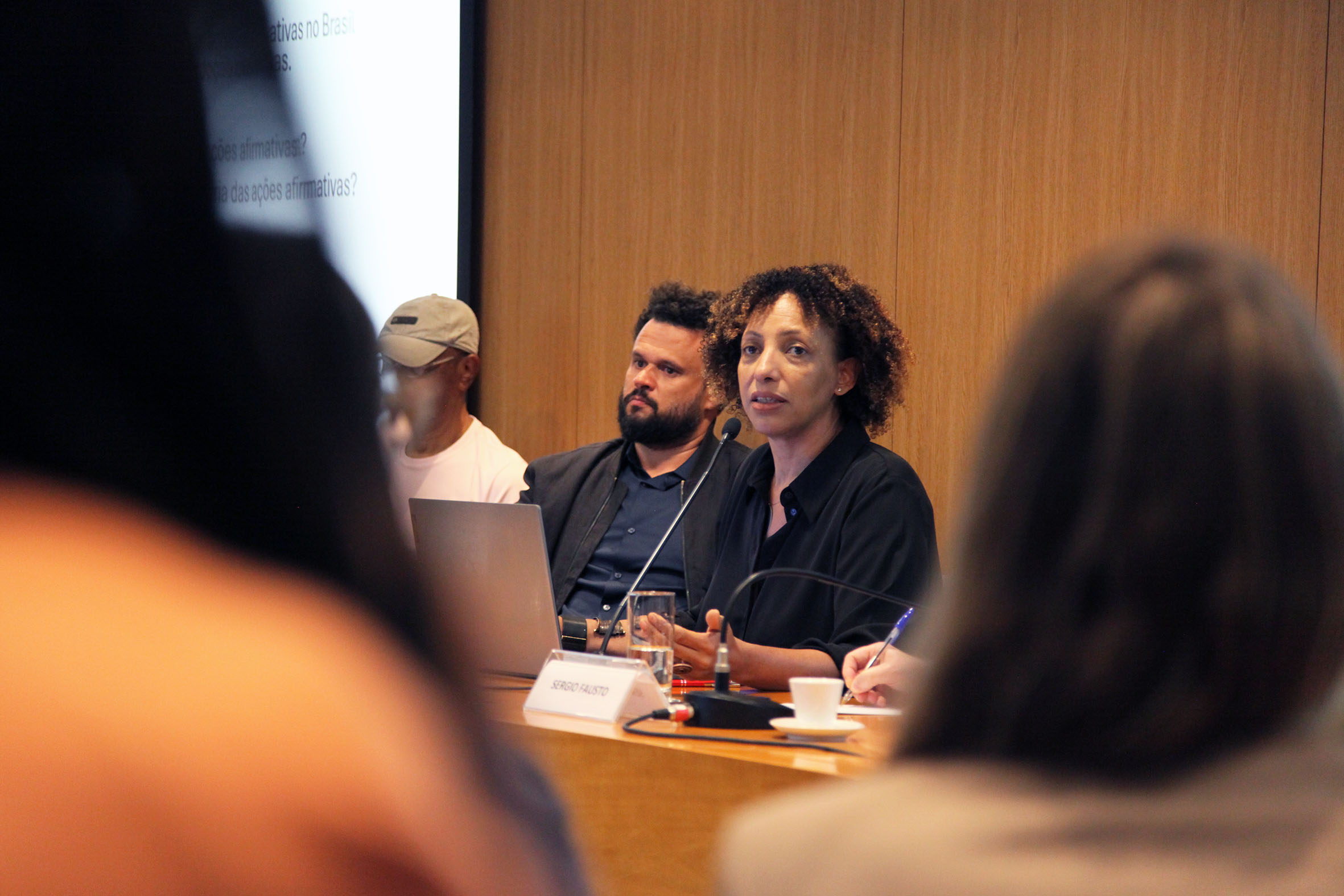 Fotografia colorida que mostra, ao centro, uma mulher e dois homens sentados atrás de uma mesa. Sobre a mesa, computadores e microfones. A mulher está falando. Atrás deles, um painel de madeira. Em primeiro plano, desfocadas, pessoas na plateia.