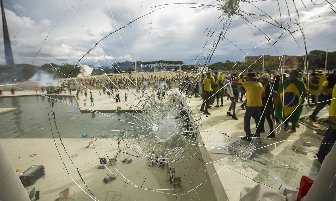Fotografia colorida que mostra a manifestação ocorrida em Brasília no dia 8 de janeiro de 2023. À direita, em pé, manifestantes. Muitos deles usam camisa verde e amarela e máscara. Em primeiro plano, um vidro trincado.
