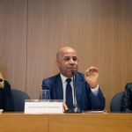 Fotografia colorida que mostra, da esquerda para a direita, Cláudia Costin, Alexsandro Santos e Raquel Teixeira. Os três estão sentados atrás de uma mesa de madeira. Alexsandro está falando e gesticulando com a mão esquerda. Cláudia e Raquel estão olhando para ele. Os três estão no auditório da Fundação FHC. Atrás deles, um painel de madeira.