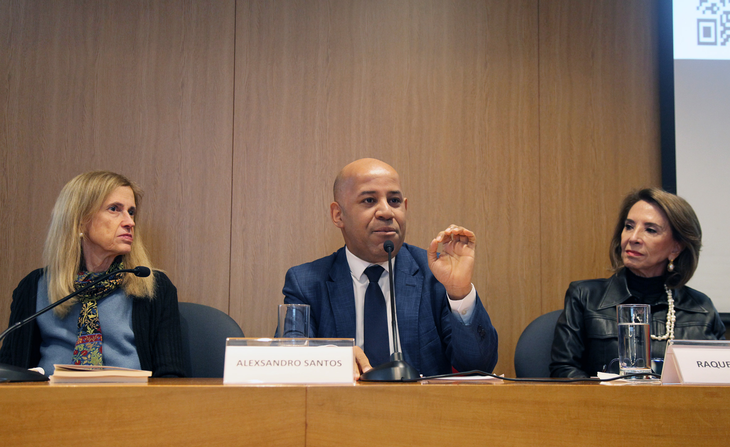Fotografia colorida que mostra, da esquerda para a direita, Cláudia Costin, Alexsandro Santos e Raquel Teixeira. Os três estão sentados atrás de uma mesa de madeira. Alexsandro está falando e gesticulando com a mão esquerda. Cláudia e Raquel estão olhando para ele. Os três estão no auditório da Fundação FHC. Atrás deles, um painel de madeira.